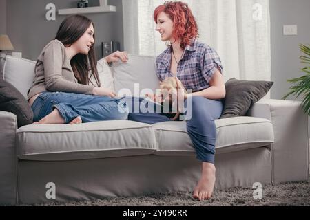 Engrossed in gossip, two young women share surprising news, their expressions vivid, with a puppy cuddled between them Stock Photo