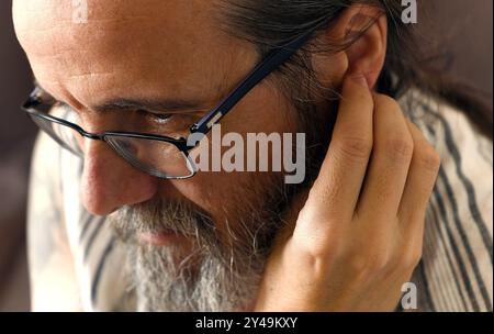 A man with glasses and a beard is touching his ear. He is looking at something with with much interest Stock Photo