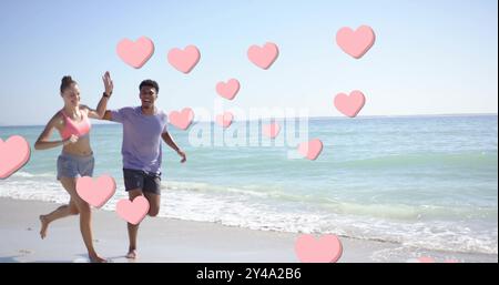 Image of pink hearts over diverse couple running on beach Stock Photo