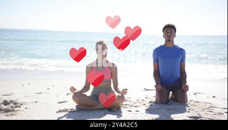 Image of red hearts over diverse couple meditating on beach Stock Photo