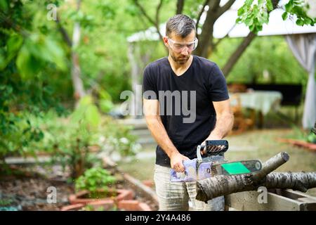 Handsome man using chainsaw for cutting tree branches at his backyard. Professional tree care Stock Photo