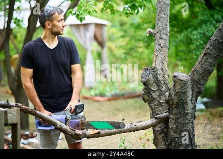 Handsome man using chainsaw for cutting tree branches at his backyard. Professional tree care Stock Photo