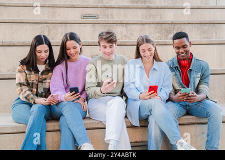 Group of multiracial young friends sitting on a staircase using their cellphones to share photos on social media. Teenage student people playing a game together with a mobile phone app on a meeting. High quality photo Stock Photo