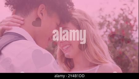 Image of hearts and pink tint over happy diverse couple embracing at outdoor wedding Stock Photo