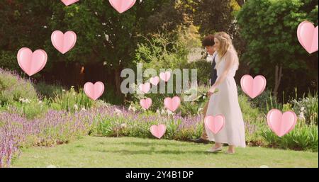 Image of pink hearts over happy diverse couple walking in sunny garden on wedding day Stock Photo