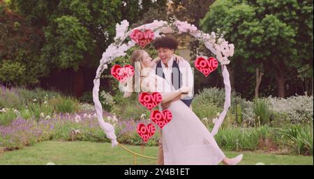 Image of red love you hearts over happy diverse couple embracing in garden on wedding day Stock Photo