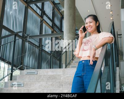An attractive, Asian woman in fashionable is talking on the phone while standing in front of the building or shopping mall. People and wireless techno Stock Photo