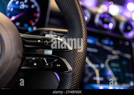 Close-up shot of a luxury car steering wheel featuring control buttons and sleek leather details, with a blurred background displaying a high-tech das Stock Photo