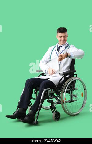 Young male doctor in wheelchair with laptop checking time on green background Stock Photo