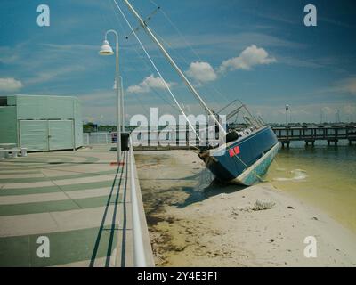 Damaged light blue and white Sailboat leaning to the right on the beach in trees after a storm in Gulfport, Florida. Looking from the beach to the bay Stock Photo