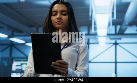 Software developer walking in data center, using tablet to design, implement and manage gear, ensuring optimal performance and reliability. IT worker in server farm coding on device, camera A Stock Photo