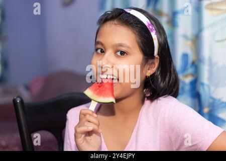 A beautiful pretty cute Indian girl child eating red watermelon with smiling face indoors Stock Photo