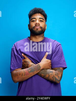 Young man in purple shirt stands confidently, crossing his tattooed arms, looking at camera with confidence against blue background. Stock Photo