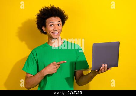 Photo of cheerful positive guy dressed green t-shirt pointing modern gadget isolated yellow color background Stock Photo