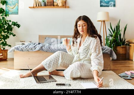 The young woman sits comfortably on the floor, sipping tea and jotting notes as she works. Stock Photo