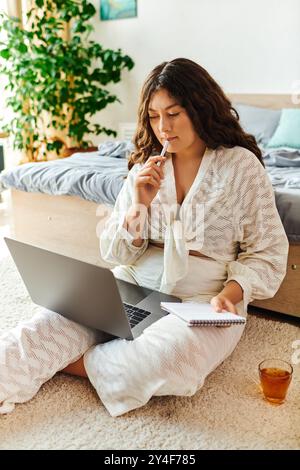 A young beautiful plus size woman thoughtfully writes in a notebook while using her laptop at home. Stock Photo