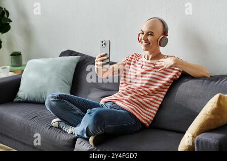 A cheerful young woman with alopecia sits comfortably on a couch, engaging in a joyful video call. Stock Photo