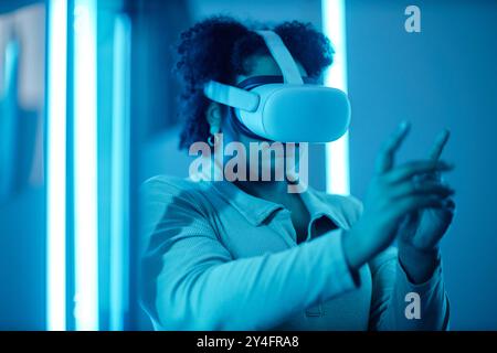 Portrait of African American woman wearing VR headset in modern art show with blue neon lights and reaching out experiencing digital cyberspace Stock Photo