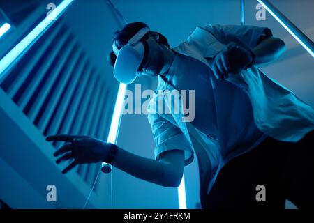 Low angle portrait of man wearing VR headset in modern art show with blue neon lights enjoying futuristic digital cyberspace Stock Photo