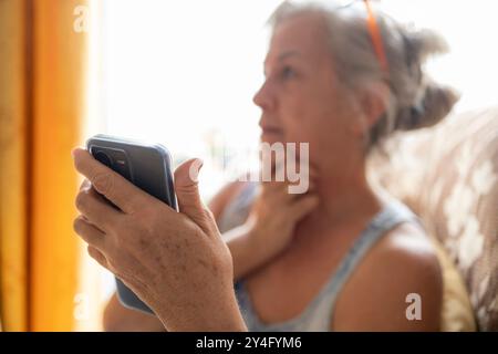 A senior woman sits at home, holding a smartphone in her hand while appearing deep in thought. Stock Photo