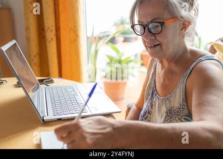A senior woman multitasks by writing notes while using a laptop at home, sitting by a bright window. Stock Photo
