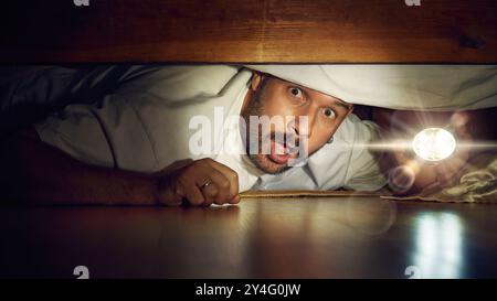 Man looking under bed with flashlight. Shocked, excited and scared face at the same time. Night time bedroom scene. Stock Photo