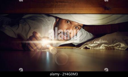 Man looking under bed with flashlight. Shocked, excited and scared face at the same time. Night time bedroom scene. Stock Photo