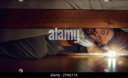 Emotional man peering under the bed with flashlight, and fearful eyes. Halloween tension, discoveries and fear Stock Photo