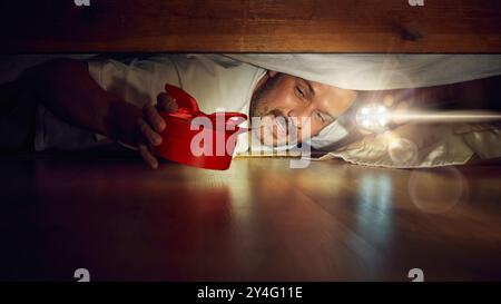 Curious and emotional man kneeling on floor, peeking under bed with flashlight. Finding hidden red present box. Stock Photo