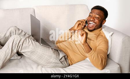 Cheerful Guy Talking On Phone Lying With Laptop On Couch Stock Photo