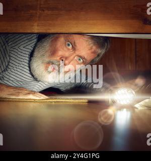 Older man peeking out from under bed, with eyes wide in surprise, looking under bed with flashlight. Spooky surprise Stock Photo