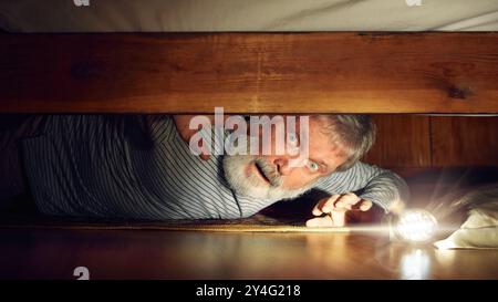 Fearful senior man looking under bed with flashlight, expressing emotions of fear, feel scared. Frightening night at home Stock Photo