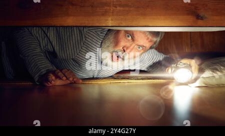 Emotional senior man looking under bed with flashlight, expressing emotions of shock. Frightening night at home. Stock Photo