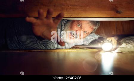 Horror. Emotional elderly man with terrifying emotions, looking under bed with flashlight, reaching out to grab Stock Photo