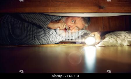 Fearful senior man looking under bed with flashlight, expressing emotions of fear, feel scared. Frightening night at home Stock Photo