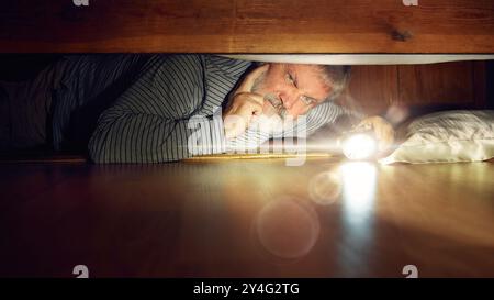 Thoughtful and emotional senior man lying on floor in bedroom, looking under bed with flashlight, searching for lost item Stock Photo