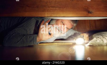 Emotional senior man lying on floor in bedroom, looking under bed with flashlight, finding lost item, looking with disgusting emotions Stock Photo