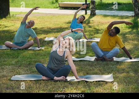 Active blond female fitness trainer in leggins and t-shirt sitting on mat with crossed legs and doing side bends in front of elderly people Stock Photo