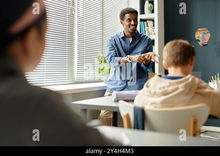 Medium shot of smiling African American male teacher talking to children in classroom with window blinds closed while counting on fingers explaining new topic to students at school, copy space Stock Photo