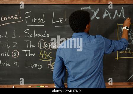 Rear view of male African American teacher writing on blackboard with piece of chalk conducting exam at primary school Stock Photo