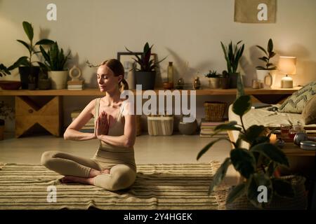 Young serene woman with namaste gesture keeping her eyes closed and legs crossed while practicing meditation exercise at home Stock Photo