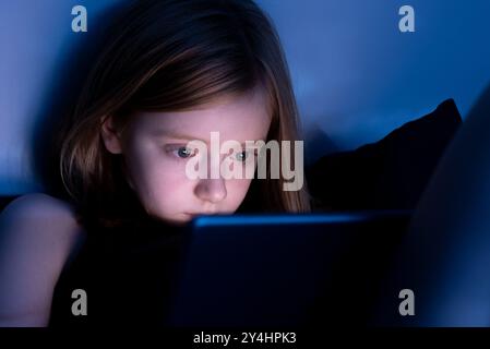 Low-light portrait of a nine year old girl using a tablet screen at night, with blue light reflecting on her face. Stock Photo