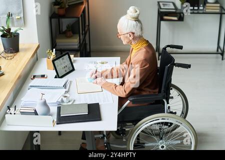 Elderly woman sitting in wheelchair while writing at desk in a modern office setting with various supplies and plants around her. Engaged in work using electronic tablet Stock Photo