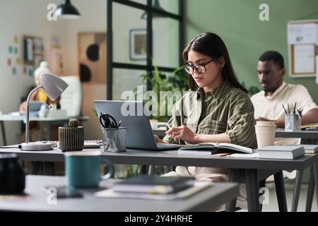 Woman in green shirt typing on laptop in modern shared workspace surrounded by colleagues, books, and plants creating collaborative environment multiple working professionals Stock Photo