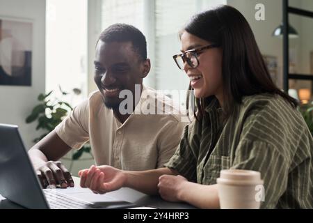 Modern office with smiling colleagues collaborating on project, both focused on laptop screen and appearing engaged in discussion Stock Photo