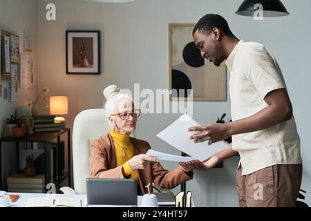 Elderly woman and young man engaged in business discussion in modern office setting, with papers, laptop, and plants around them. Background has creative elements and professional ambiance Stock Photo