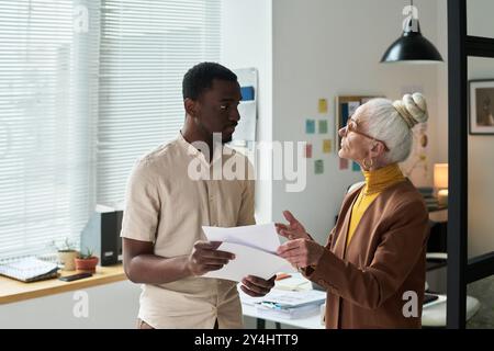 Senior woman and young man discussing project details in modern office. Both individuals are focused on conversation while standing near workspace wit Stock Photo