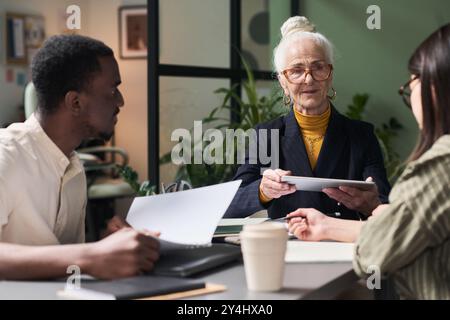Group of diverse colleagues discussing project details around meeting table, with senior woman holding tablet and contributing to conversation Stock Photo