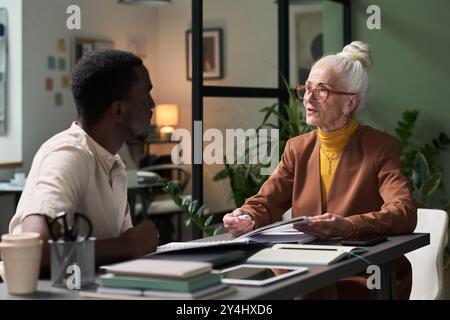 Senior woman with glasses sitting at desk, going over business documents with young male colleague in modern office setting, plants and office supplies background Stock Photo