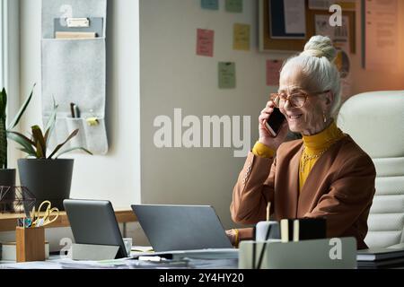Senior woman sitting at desk, smiling while talking on phone and looking at laptop. Office setting includes files, sticky notes, and indoor plants for Stock Photo